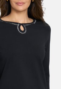 Top negro de manga larga con escote redondo, que presenta un corte en forma de llave y acentos metálicos a lo largo del cuello. Textura suave.