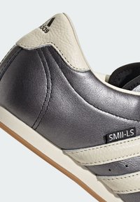 Szare skórzane sneakersy o teksturowanej powierzchni, z kremowymi akcentami i trzema białymi paskami. Posiadają beżową gumową podeszwę i metkę z logo na pięcie.
