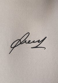 Firma in ricamo nero su tessuto beige chiaro e strutturato, caratterizzata da lettere corsive fluide con spessori variabili.