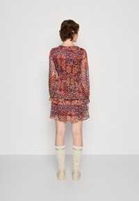 Robe transparente à motifs dans des tons chauds de rouge et de bleu, avec des manches longues, un ourlet en couches, et accompagnée de bottes beiges montantes.