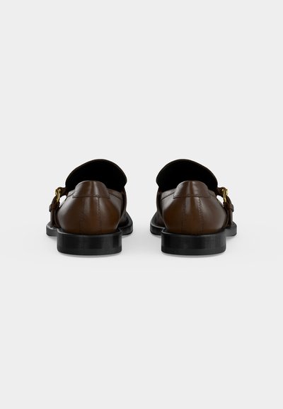Paire de mocassins en cuir brun foncé avec semelles noires et boucles dorées, vus de dos sur un fond clair uni.