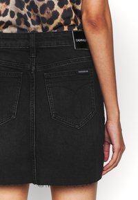 Person som bär en svart denimkjol från Calvin Klein med bakfickor och en leopardmönstrad topp, med en avslappnad hand vid sidan mot vit bakgrund.