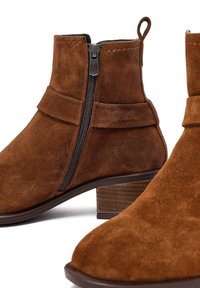 Stivali alla caviglia in suede marrone con zip laterale, bordo superiore curvo e tacco basso impilato. Presentano una texture liscia e dettagli di cucitura minimi.