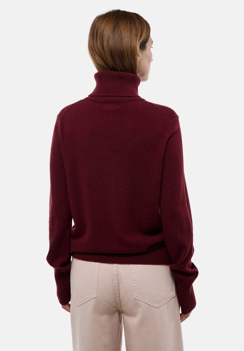 Pull à col roulé bordeaux en tissu doux, avec un col côtelé et des manches longues. Porté avec un pantalon ajusté beige clair.