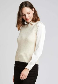 Pull à sans manches de couleur crème clair avec un ourlet côtelé, associé à une chemise blanche à manches longues avec col. Fil texturé avec un léger éclat.