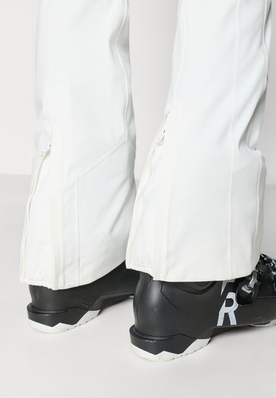 Bela smučarska hlače z zadrgami pri gležnjih, združene s črnimi smučarskimi čevlji z sivimi poudarki in teksturirano površino.