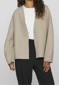 Ljus beige ullkofta med lös passform, breda ärmar och två framknappar. Bärs över en vit skjorta och matchas med svarta byxor.