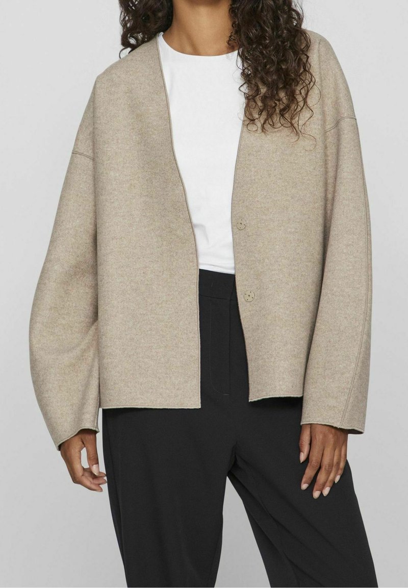 Ljus beige ullkofta med lös passform, breda ärmar och två framknappar. Bärs över en vit skjorta och matchas med svarta byxor.