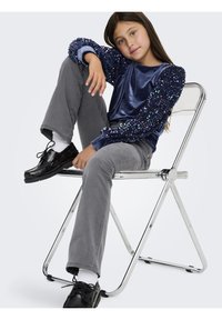 Camisa de veludo azul-marinho com mangas bufantes com lantejoulas, calças cinzentas e sapatos pretos. A criança está sentada casualmente numa cadeira dobrável cromada.