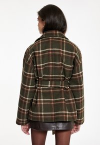 Mujer con cabello oscuro que lleva una chaqueta de cuadros verde oliva y marrón con cinturón, sobre una falda de cuero marrón oscuro, de espaldas sobre un fondo blanco.