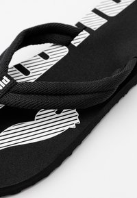 Czarne klapki z teksturowaną podeszwą, z paskiem z białym, wąskim wzorem logo oraz plecionymi czarnymi paskami. Płaski, prosty kształt idealny do codziennego noszenia.