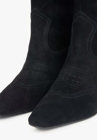 Černé semišové kotníkové boty s špičatou špičkou, vyznačující se zdobeným prošíváním na svršku a zakřivenými výřezmi na stranách.