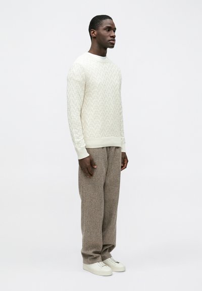 Pull texturé crème clair avec encolure ronde, associé à un pantalon ample en tweed marron et des baskets blanches, debout devant un fond uni.