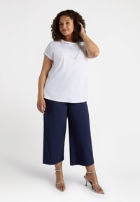 Vit kortärmad topp med silverkedjeaccent, tillsammans med mörkblå vida byxor och silverfärgade remsandaler, på en ljus bakgrund.