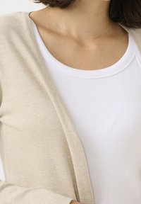 Kobieta w białej prążkowanej koszulce na ramiączkach pod beżowym kardiganem, odsłaniająca ramię i górną część klatki piersiowej.