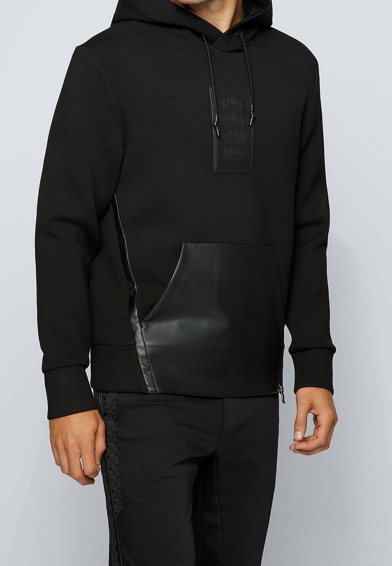 Homme portant un sweat à capuche noir avec un écusson logo en relief et une poche avant ainsi que des détails latéraux en texture cuir, assorti à un pantalon noir.