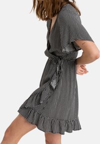 Nainen pukeutuneena mustavalkoiseen ruudulliseen lyhythihaiseen kietaisumekkoon, jossa on röyhelöhelma, seisoo dynaamisessa asennossa yksinkertaista taustaa vasten.