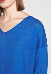 Pull léger bleu avec décolleté en V, coupe ample et manches courtes. La texture semble douce et légèrement transparente, avec des détails de couture subtils.