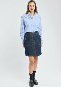 Jupe en denim en forme de A avec deux poches avant et des accents de boutons dorés, associée à une chemise à boutons rayée verticalement de couleur bleu clair.