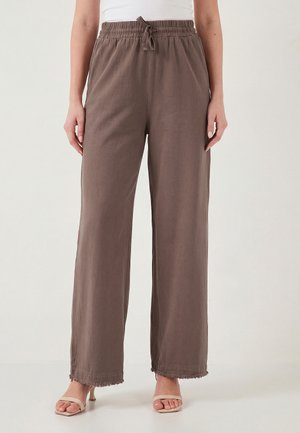 Inimene, kes kannab lahtisi pruune pükse elastse nöörvöökohaga ja narmendavate äärtega, kombineerituna beežide avatud varbaga kontsakingadega.