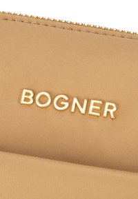Béžová látková peněženka se zlatým logem "BOGNER". Povrch má texturovaný vzor a zipový detail podél horního okraje.