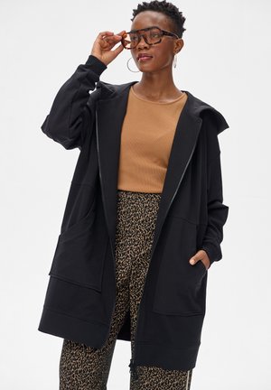 Vrouw met een oversized zwarte hoodie met rits, een bruin topje, een broek met luipaardprint, grote bril met tortoise montuur en oorringen, poseert met een hand op haar bril.