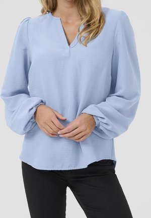 Femme portant un chemisier bleu clair à manches longues bouffantes et un pantalon noir, mains doucement jointes au niveau de la taille.