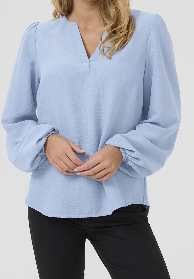 Femme portant un chemisier bleu clair à manches longues bouffantes et un pantalon noir, mains doucement jointes au niveau de la taille.
