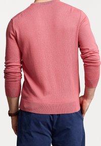 Rosa stickad tröja med långa ärmar, rund hals och ribbade muddar och fåll. Tyget har en jämn struktur. Bakåtsidan visar passformen mot kroppen.