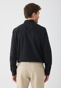 Mann, der ein langärmliges schwarzes Hemd und beige Hose trägt, steht mit dem Rücken zur Kamera vor einem schlichten weißen Hintergrund.