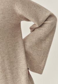 Beige kötött pulcsi ujja, amely lazán illeszkedik, puha textúrájú és bordás mandzsettával rendelkezik, részben betűrve a részletek kiemelése érdekében.