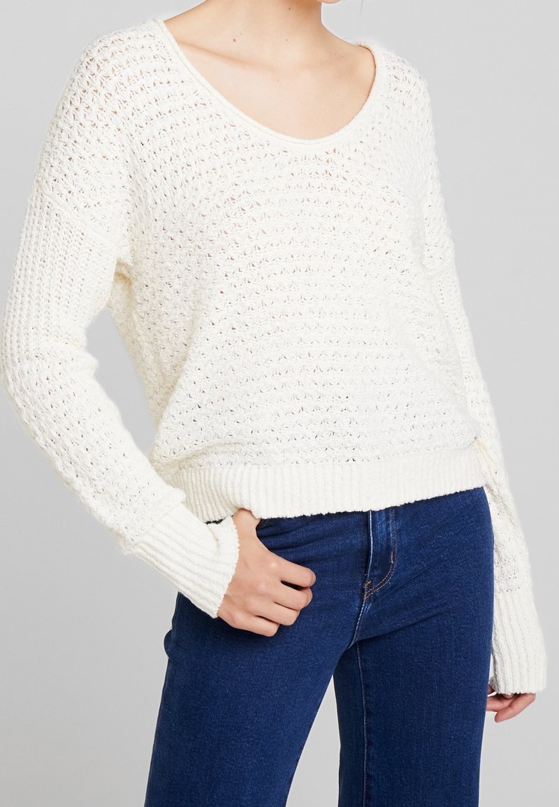 Biały sweter z dzianiny o luźnym kroju, z dekoltem w kształcie litery V, prążkowanymi mankietami i dołem oraz teksturowanym, otwartym wzorem oczek. Połączony z niebieskimi dżinsami.