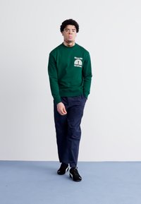 Groene sweatshirt met een logo op de borst, gecombineerd met marineblauwe broek en zwarte sportschoenen, staand op een lichtblauwe vloer.
