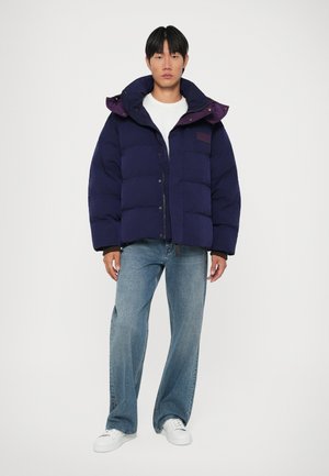 Giubbotto imbottito blu navy con colletto alto e dettagli viola, dotato di chiusura con zip, design trapuntato e due tasche laterali. Indossato con jeans blu.