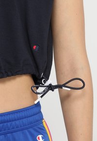 Černé zkrácené tričko vyrobené z bavlny s utažitelnou šňůrkou na spodním lemu. Obsahuje malé vyšívané logo na boku.