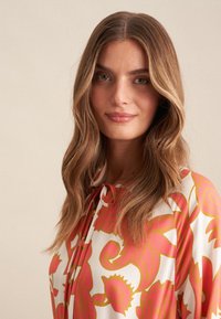 Langärmlige Bluse in Weiß mit großem rosa und gelbem Blumenmuster. Mit einer Bindeschließe am Hals und einer lockeren Passform.