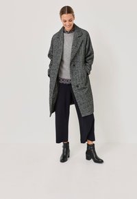 Pelēks teksturēts garais mētis ar plašu apkakli, kas kārtota virs pelēka džempera un rakstainas krekla, apvienots ar tumšām biksēm un melniem potīšu zābakiem.