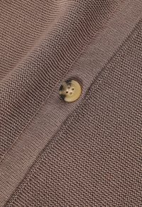 Brązowa dzianina o fakturze małych, równomiernych ściegów. Na pierwszym planie znajduje się okrągły, jasny guzik z czterema otworami.