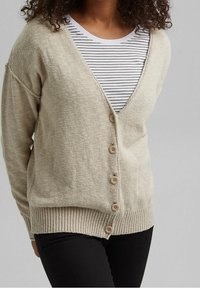 Cardigan beige a maglia con scollo a V, cinque bottoni e orlo a costine; indossato sopra una maglietta a righe, con maniche leggermente oversize.