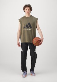 Young man in sleeveless olive basketball shirt holding a basketball, wearing black pants with white accents and purple sneakers.