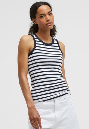 Frau mit lockigem Haar, die ein ärmelloses weißes Tanktop mit marineblauen horizontalen Streifen und weiße Hosen vor einem einfarbigen hellen Hintergrund trägt.