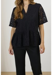 Top in pizzo nero con scollatura a V, maniche corte e orlo a peplo svasato. Motivo floreale intricato su tutta la superficie. Abbinato a pantaloni neri.