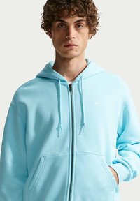 Νεαρός άνδρας με κατσαρά μαλλιά φοράει μια ανοιχτό γαλάζια μπλούζα με φερμουάρ Nike SB, με το ένα χέρι στην τσέπη, σε απλό ανοιχτό φόντο.