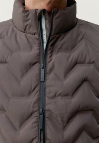 Bruine gewatteerde jas met een hoge kraag, voorzien van een zwarte rits en een zigzag stikselpatroon voor extra textuur en ontwerpdetails.