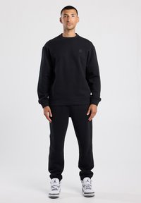 Felpa nera e pantaloni coordinati, realizzati in tessuto morbido. Presentano un collo a giro e polsini a costine. Indossati con sneaker bianche.