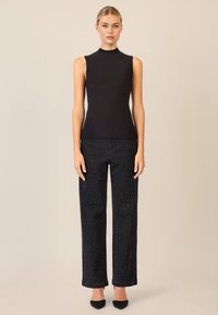 Top smanicato nero con colletto alto, abbinato a jeans scuri a vita alta con piccole borchie bianche e taglio dritto.