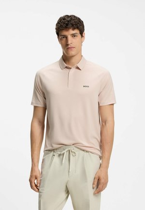Mężczyzna w jasnobeżowej koszulce polo z logo „BOSS” i jasnobeżowych spodniach z sznurkiem, stojący na białym tle.