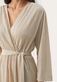 Wickeldress in hellem Beige mit vertikaler Ripptextur, langen Ärmeln, tiefem V-Ausschnitt und einem gebundenen Taillengürtel. Weicher Stoff mit subtiler Glanz.