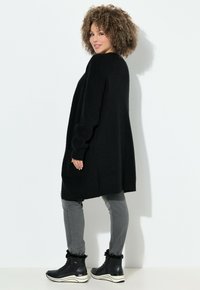 Kvinna med lockigt hår iförd en lång svart kofta, grå jeans och svarta stövlar med pälsfoder, stående mot en vit vägg.