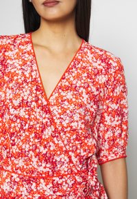 Robe portefeuille rouge à fleurs avec des manches courtes, présentant un décolleté en V profond, des finitions orange contrastantes et un accent noué à la taille.